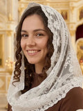 A smiling woman wearing a white Italian bridal lace-trimmed veil over her head and shoulders, paired with a brown dress, set against a softly blurred ornate interior background. 