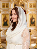 A woman wearing a white traditional Orthodox dress with a sheer white veil (khimar) decorated with pearl-like beads along the edge, standing in a golden, ornate church interior with religious icons.
