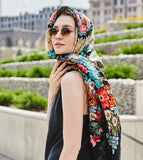 A woman cozying up in a very large winter shawl made from natural merino wool worn over her head, highlighted by vivid pink peonies, blue wildflowers with yellow centers, and a dramatic black fringed hem.