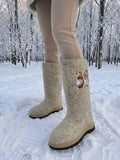 A person stands in a snow-covered forest wearing fashionable Slavic felt valenki boots featuring a long, wide shaft that comfortably accommodates tucked-in beige leggings. The boots are made from light oatmeal-colored melange wool felt, adorned with a large, elegant embroidery of a brown and white flower with delicate swirling branch details on the outer side. The bottom features a modern, thick rubber outsole with a black grooved tread and a light tan midsole layer for walking on winter snow.