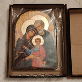 A framed religious icon depicting the Holy Family, still in its outer cardboard packaging, placed on a quilted white fabric surface. Domestic prayer corner setup showcasing the Holy Family wood-backed icon standing upright. The rich colors and striking gold leaf pattern create a spiritual focal point for family devotion.