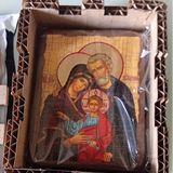 A religious icon painting of the Holy Family, still sealed in its clear plastic protective wrapping and presented inside a brown cardboard shipping box on a light-colored surface.