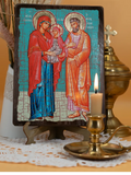 A rectangular religious icon plaque showing the Holy Family (Mary, Joseph, and baby Jesus) on a dark wooden frame, displayed on a small stand with a lit candle and brass vessel in front, against a warm beige background. 