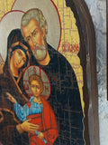 A close-up of a wooden Orthodox Christian icon depicting the Holy Family (Joseph, Mary, and baby Jesus) on a white textured wall next to another icon.