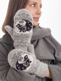A close-up of a woman wearing gray knit mittens with black horse and white winter patterns, paired with a gray coat and scarf against a plain white background.