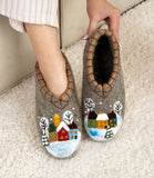 Pair of woolen slippers with winter scene design held by a hand on a carpeted floor.