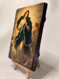 Devotional Marian artwork of the Blessed Virgin Mary untying a white ribbon surrounded by angels. Close-up of a rustic wood block icon featuring Mary Undoer of Knots with gold leaf details. 