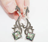 Ornate silver earrings with pale green gemstones and marcasite held by a hand against a white background