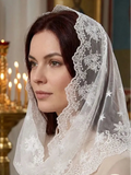 A woman wearing a white lace-trimmed veil with embroidered floral and star motifs, posed indoors in front of a softly blurred candelabra.