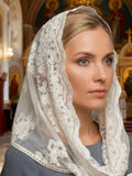 A woman wearing a white lace Orthodox veil (pikhra) over her head and shoulders, set against the background of a decorated church interior. 