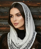 A woman wearing a white sheer tulle italian style bridal head cover with white embroidered dots and scalloped edges, posed against a rustic dark wood wall.