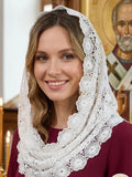 A smiling woman wearing a white lace Slavic head covering in a church setting with a saint icon and candles in the background. 