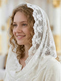 A woman wearing a white lace-trimmed veil and cream-colored dress, smiling in a softly blurred indoor setting with warm golden light.