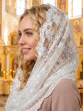 A woman wearing a white embroidered lace head covering or veil, photographed indoors in a brightly lit church with gold-toned iconostats in the background.