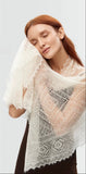 A woman comfortably wrapping herself in a warm, lace-detailed winter pashmina shawl in ivory. The delicate texture of a gossamer, knitted ecru pashmina shawl worn as a shoulder wrap.