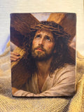A rectangular wooden icon featuring a painted portrait of Jesus carrying a wooden cross, displayed on a textured beige burlap fabric. Handcrafted wooden icon of Jesus Christ carrying the heavy wooden Cross on His shoulder.
