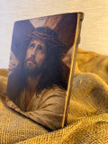 A rectangular decorative wall art icon  featuring a painted portrait of Jesus with a crown of thorns, standing upright on a burlap fabric surface against a light wall. Realistic portrait painting of Jesus with the Crown of Thorns printed on a thick wood block.