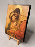 Sacred Christian artwork of the Holy Family in warm golden and red tones for home altars. Close-up of a handcrafted wood icon depicting the nativity family with a textured gesso finish.