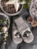 A pair of gray knit winter mittens with black horse motifs and snowflake details, styled in a cozy flat-lay arrangement with pinecones, a star ornament, and a gray knit scarf on a dark gray fabric background.