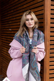 Woman wearing a pink coat and patterned merino wool gray scarf against a wooden background. Close-up view of a premium merino wool scarf, highlighting the woven gray and ivory geometric Slavic motifs accented with deep blue tones.