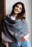 Woman wearing a gray and blue patterned merino wool shawl with a wooden door and white wall background. A tailored dress form displaying an oversized blanket-style shawl crafted from merino wool, featuring a traditional Slavic pattern in a calm palette of gray, ivory, and blue.
