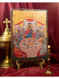 A wooden Orthodox Christian icon of the Last Supper on a small wooden stand, displayed on a red satin drape with a brass chalice and a lit candle. Mystical Supper wooden icon of Jesus Christ and the Apostles on a desk with a burning candle.