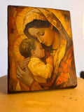 A rectangular religious art piece depicting the Madonna and child in a warm, textured mosaic style, leaning against a light background on a wooden surface. Close-up of a contemporary Catholic Mary and Jesus icon featuring textured faux-gesso finish.