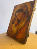 A small rectangular wooden icon featuring a painted religious scene of a woman cradling a child, photographed standing on a light wood surface against a plain white wall. Modern Christian home decor icon of the Blessed Mother and Christ Child with gold gilding.