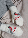 A pair of white knit sheep wool mittens with a hand-knitted robin design in red, black, and gray, resting on a beige and pink herringbone blanket next to gray knit socks. White wool mittens featuring a detailed red bullfinch bird and rowan berries design. Comfortable knit mittens in pristine white color made from natural wool and polyester.