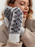 A close-up of a person wearing cream knit sheep wool mittens with a dark gray Nordic snowflake pattern, paired with a beige turtleneck sweater.