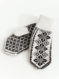 A pair of knit sheep wool mittens in black and white, featuring a black and white houndstooth pattern on one mitten and a black geometric snowflake pattern on the other, with a ribbed white cuff.