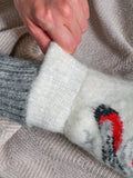 A close-up of a person's hand pulling up the cuff of a thick, fuzzy knit mitten. The mitten is primarily off-white with a gray ribbed cuff and a red and black knit pattern. Fluffy winter mittens in snow white color with traditional bird and berry embroidery. Elegant winter themed apparel showing fluffy white mittens with a festive holiday design.