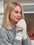 A woman wearing a gray knit sweater holds her hands together wearing white knit woolen mittens with a red and black snowman design, set against a cozy indoor background with a fireplace. Cozy 80 percent wool mittens for women designed for freezing cold winter weather. Thick and wind resistant snowy white mittens ideal for outdoor winter activities.