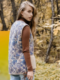 A woman stands on a leaf-strewn outdoor path wearing a sleeveless patterned woolen vest over a brown long-sleeve top, light blue jeans, and white sneakers, with a white dog beside her. 
