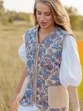 A woman walking outdoors in a grassy field wearing a blue floral embroidered vest over a white puff-sleeve dress, holding a woven basket. 