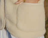 Close-up of a cream-colored sheepskin vest with a large front patch pocket, worn by a person whose hand is tucked inside the pocket. 
