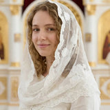 A woman wearing a white embroidered lace head covering and matching white dress, standing in front of a softly blurred ornate interior with gold and cream church architectural details.