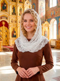 A smiling blonde woman wearing a brown dress and a white lace-trimmed head covering stands inside a brightly lit Orthodox church with ornate gold iconostats and religious paintings in the background.