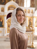 A smiling young woman in a brown habit and white lace head covering stands inside a bright, ornate church with gold and white architectural details.