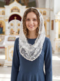 A smiling woman with curly light brown hair wears a sheer white lace head covering and a navy blue dress, standing in front of a softly blurred ornate church interior.