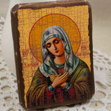 A rectangular religious icon plaque depicting the Virgin Mary with a gold background and brown border, placed on a white lace doily with a soft white backdrop.