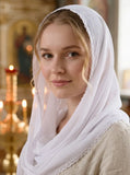 A woman wearing a white sheer headscarf with delicate lace trim, styled in a softly lit indoor setting with blurred candles and warm background tones.
