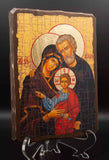 A traditional wooden Christian icon of the Holy Family (Mary, Joseph, and Jesus) displayed on a small wooden stand. The icon is styled with a lit candle in a brass holder, a wooden rosary, and a small wooden cross on a white lace doily, with dried flowers in the background.