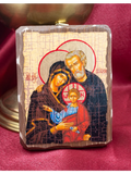 A rectangular wooden Christian icon plaque depicting the Holy Family (Mary, Joseph, and Jesus) with a textured cracked wood finish, displayed on a deep red satin fabric background covering the altar.