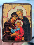 A hand holds a small wooden Orthodox icon depicting the Holy Family (Mary, Joseph, and baby Jesus) with a cracked, distressed finish. The background is a blurry outdoor setting.