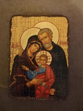 A religious icon painting of the Holy Family on a rustic, distressed wooden panel, displayed against a dark textured fabric background.