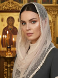 A woman wearing a white sheer Orthodox veil with white polka dots and a scalloped lace border, styled over her head and shoulders. She is wearing a dark gray habit in front of a warm, ornate church interior with gold icons and lit candles.