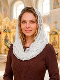 A woman wearing a white lace head covering and a brown embroidered top, standing in a brightly lit church interior with ornate gold-toned iconography in the background.