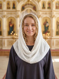 A smiling woman with blonde curly hair wearing a white lace-trimmed head covering over a dark navy dress, standing in front of a softly blurred ornate church interior with gold-toned arches and religious icons.