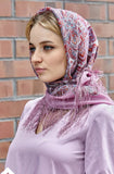 Woman wearing a patterned headscarf and light purple dress against a brick wall.